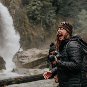 Woman smiles while holding a camera. A waterfall is in the background