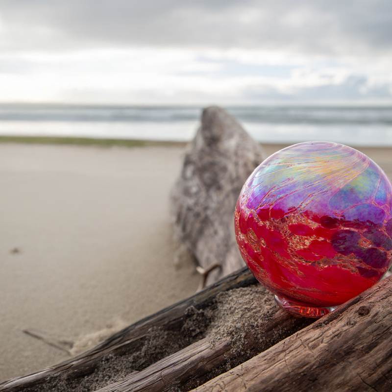 Red and purple glass ball resting on driftwood along the shore in Lincoln City, Oregon