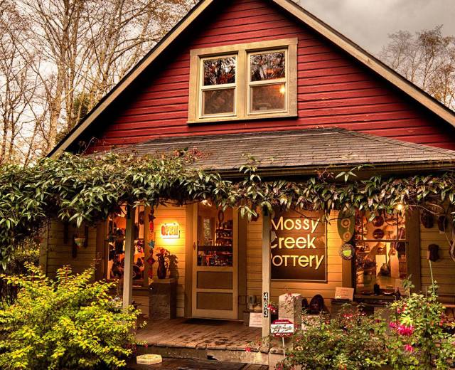 Red house with a covered porch surrounded by plants and a sign reading &quot;Mossy Creek Pottery&quot;