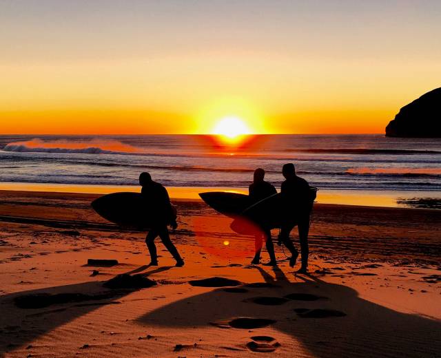 three people carrying surfboards on the beach silhouetted by the setting sun